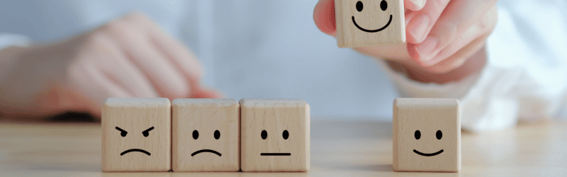 Wooden Blocks with Facial Expressions in a Row and Smily Face Block Being Picked Up by Human Hand in the Background